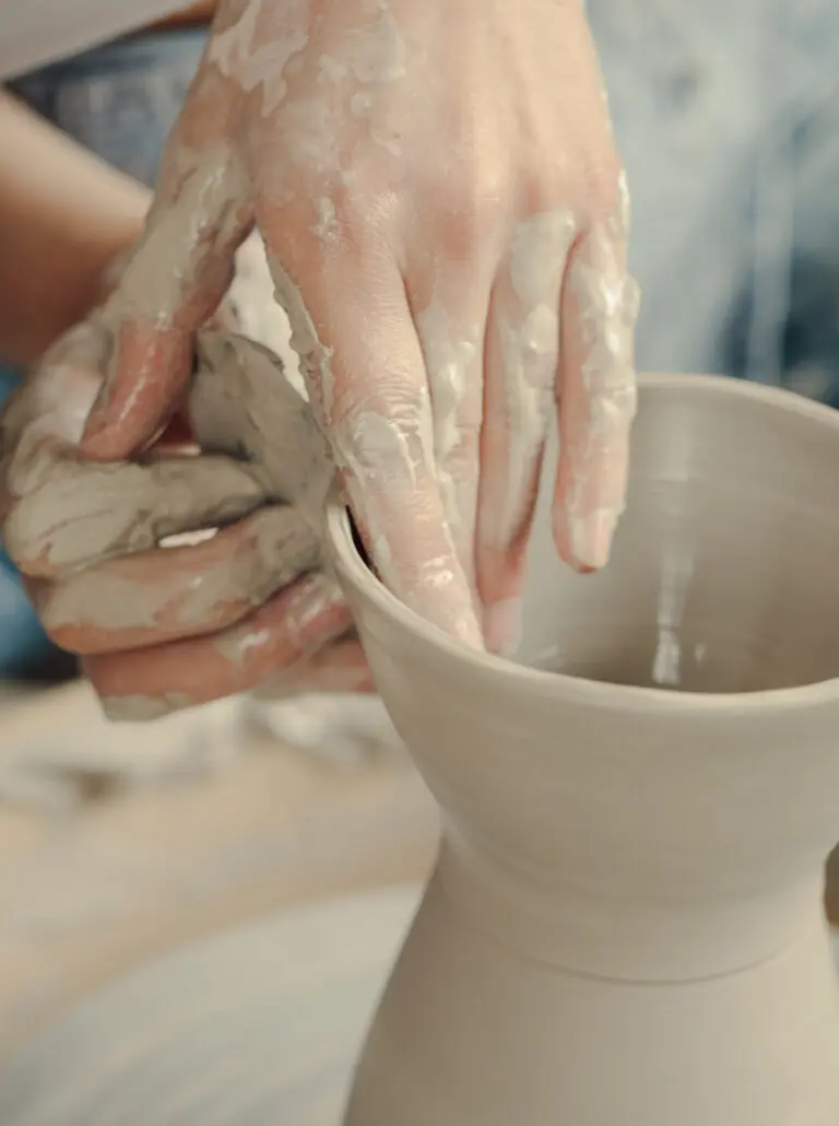 A close-up photograph of two hands working clay into a vase-like sculpture on a ceramics wheel.