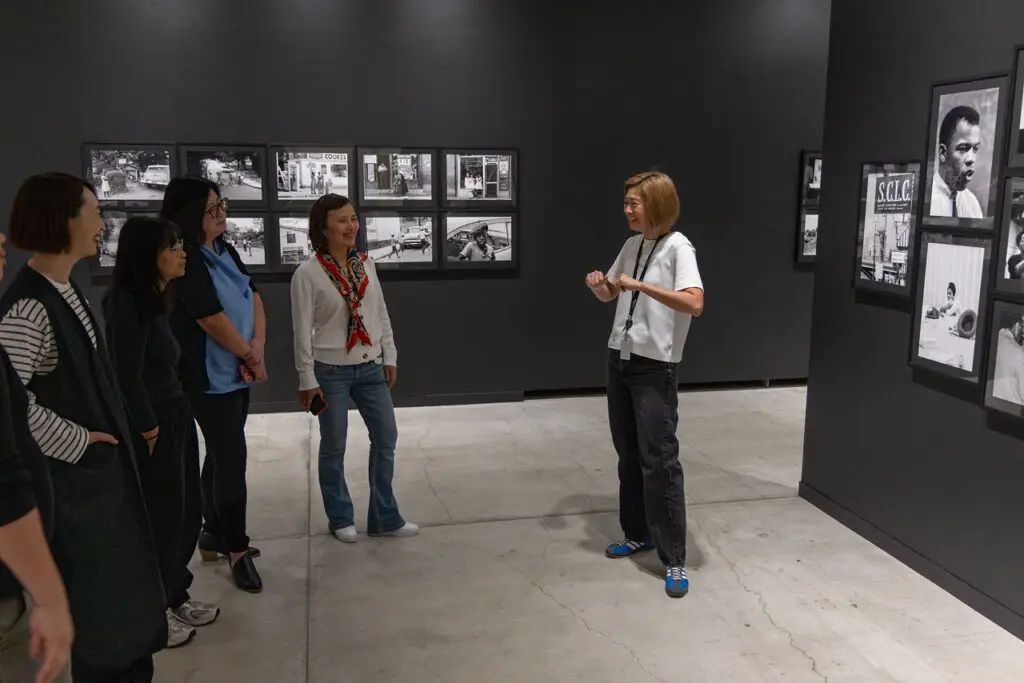 A photograph of a Gallery Tour of the exhibition "Enemy Alien: Tamio Wakayama." The photograph shows a group of Asian women from the side, smiling and engaging, while looking at a female tour guide, who is discussing the black-and-white photographs on the black wall behind her.