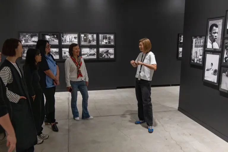 A photograph of a Gallery Tour of the exhibition "Enemy Alien: Tamio Wakayama." The photograph shows a group of Asian women from the side, smiling and engaging, while looking at a female tour guide, who is discussing the black-and-white photographs on the black wall behind her.