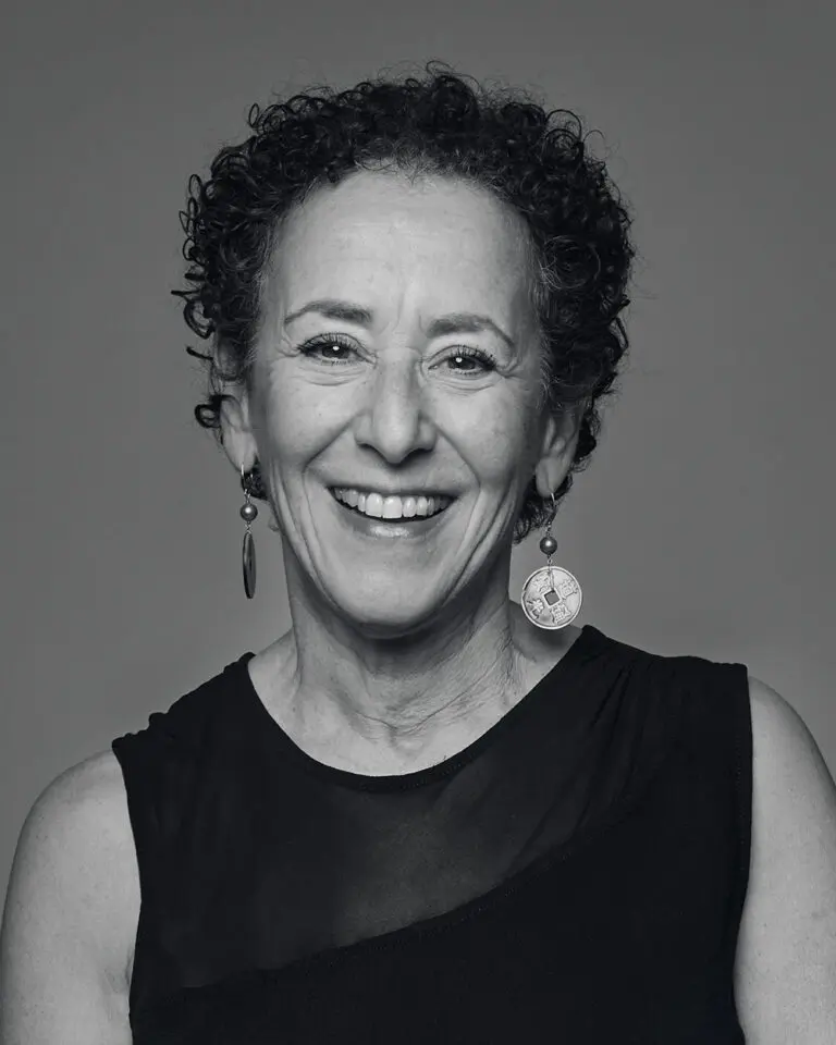 A black-and-white headshot of a woman with short brown, curly hair, standing facing the camera and wearing a short sleeved shirt. She is wearing dangling earrings.