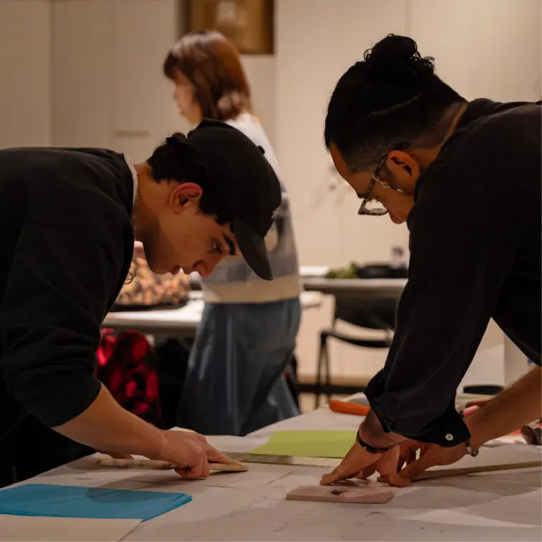 A square photograph of a teen boy wearing a hat bent over a table working on a project on the left and a man wearing glasses and with his hair in a bun leaning over the other side doing the same.
