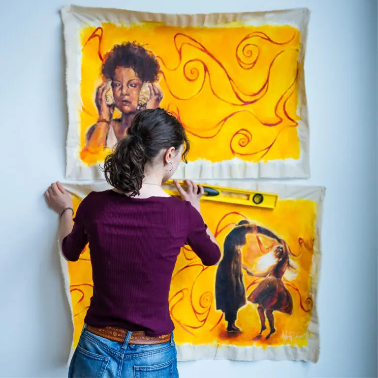 A square photograph of a teen girl standing and installing bright yellow canvas artworks on a white wall in front of her.