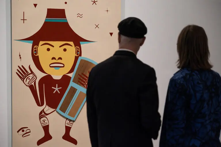 A photograph of two people, a man and a woman, from behind, looking at a painting by Bracken Hanuse Corlett of a man waving and holding a shield.