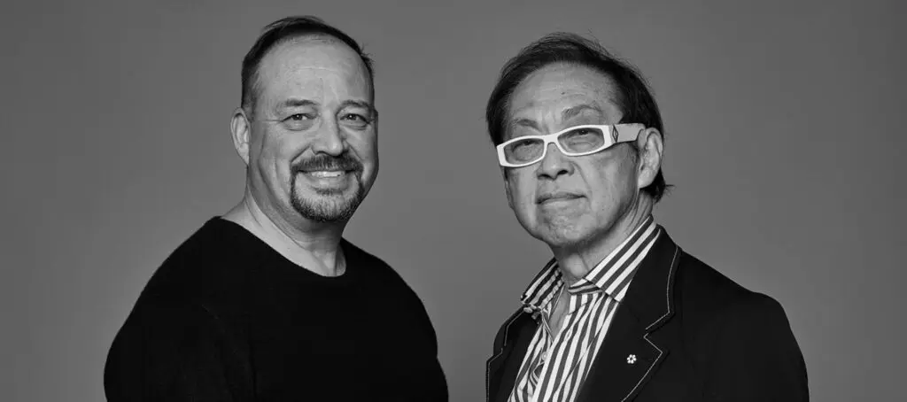 Black-and-white photograph of two men from the chest up, standing against a grey background. On the left is Alfred Waugh, an Indigenous man, wearing a black shirt. He looks out at the camera smiling. On the right is Bruce Kuwabara, a Japanese Canadian man, wearing light coloured, thick-framed glasses, a black blazer, and a button-up shirt.