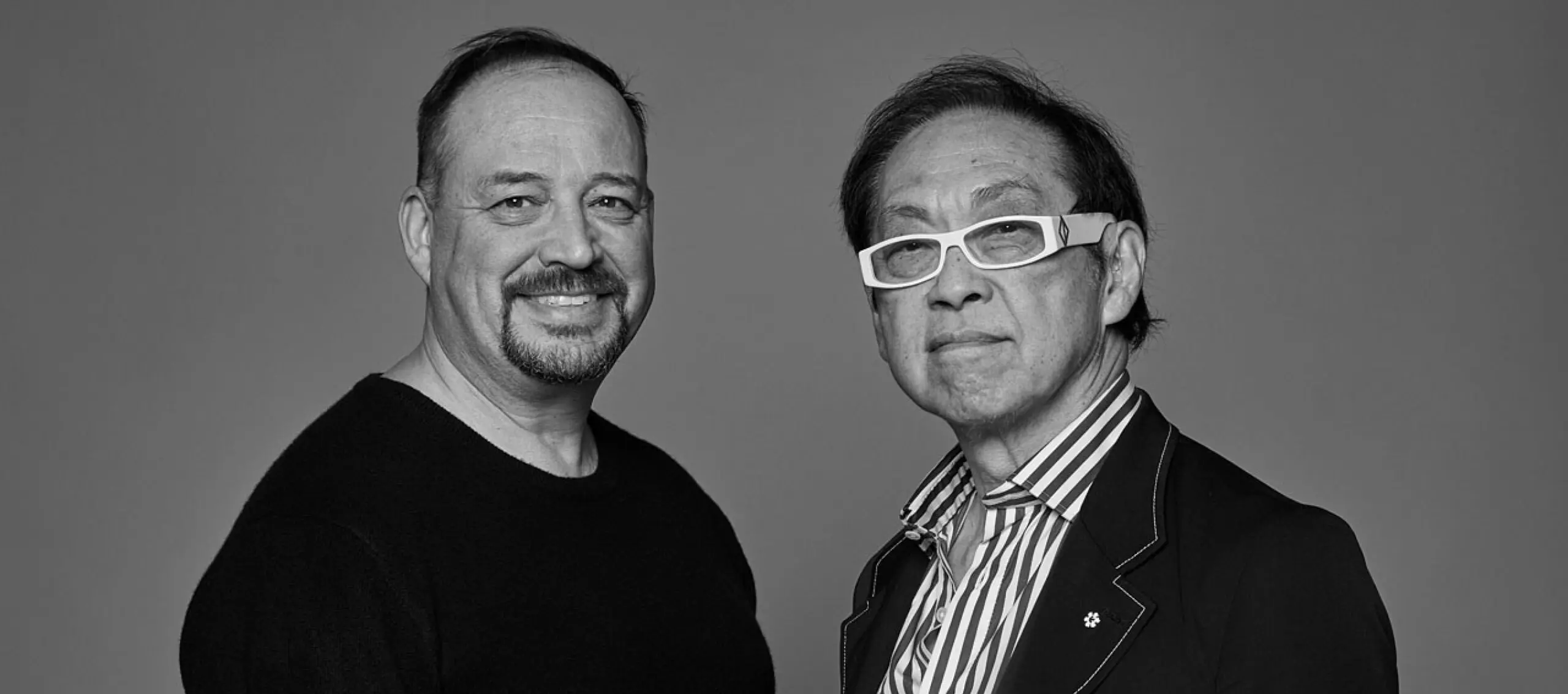 Black-and-white photograph of two men from the chest up, standing against a grey background. On the left is Alfred Waugh, an Indigenous man, wearing a black shirt. He looks out at the camera smiling. On the right is Bruce Kuwabara, a Japanese Canadian man, wearing light coloured, thick-framed glasses, a black blazer, and a button-up shirt.