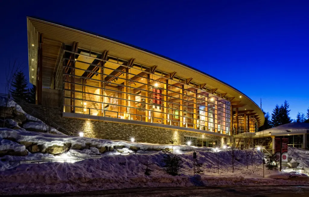 Photograph of a building at dusk. The wood building resembles a longhouse and has large windows, through which light emanates