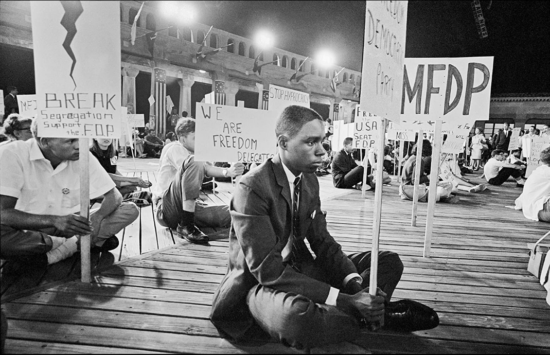 A film still from the documentary Freedom Summer of a photograph of a Black man sitting at during a demonstration. He is surrounded by other protesters holding signs.