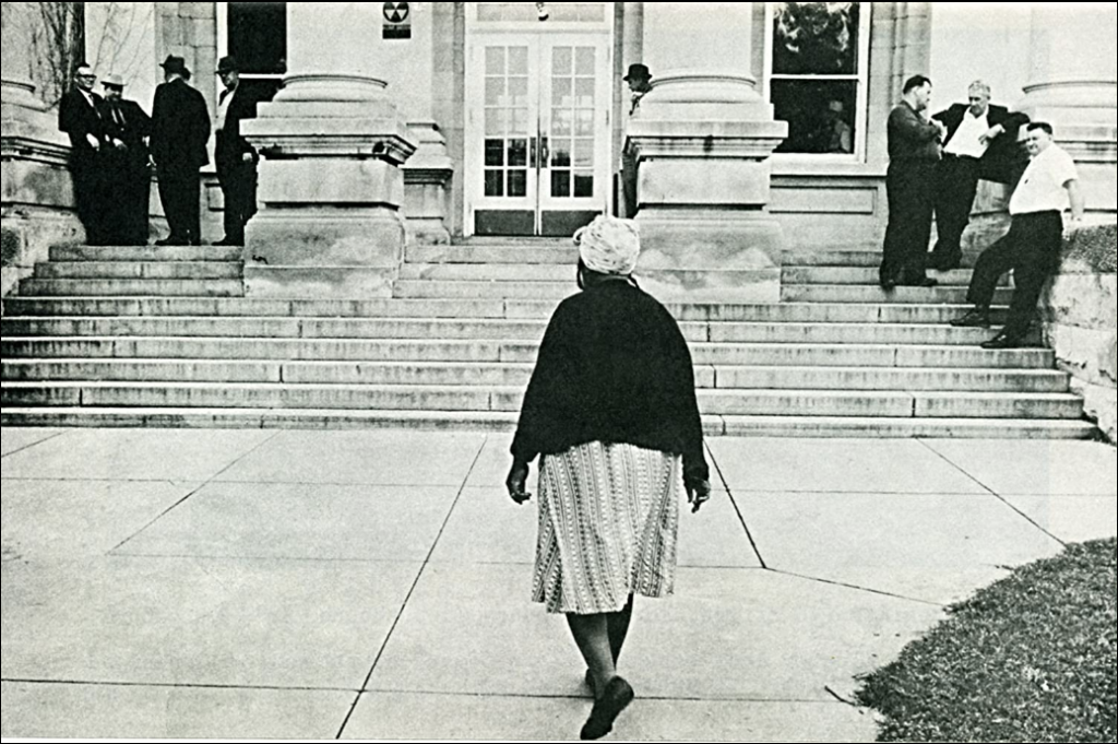 A film still from the documentary Freedom Summer of a black-and-white photograph of a woman walking up to the steps of a building, where white men stand on either side.