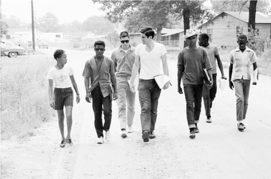A film still from the documentary Freedom Summer of a black-and-white photograph of 7 young men–2 white in the middle and 5 Black–walking down a dusty road.