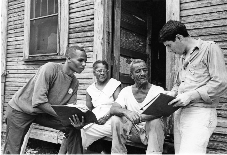 A film still from the documentary Freedom Summer of a black-and-white photograph of 3 African Americans–a young man, an older woman wearing glasses, and an older man–sitting on their porch and speaking to a young white man, who is showing them a book.