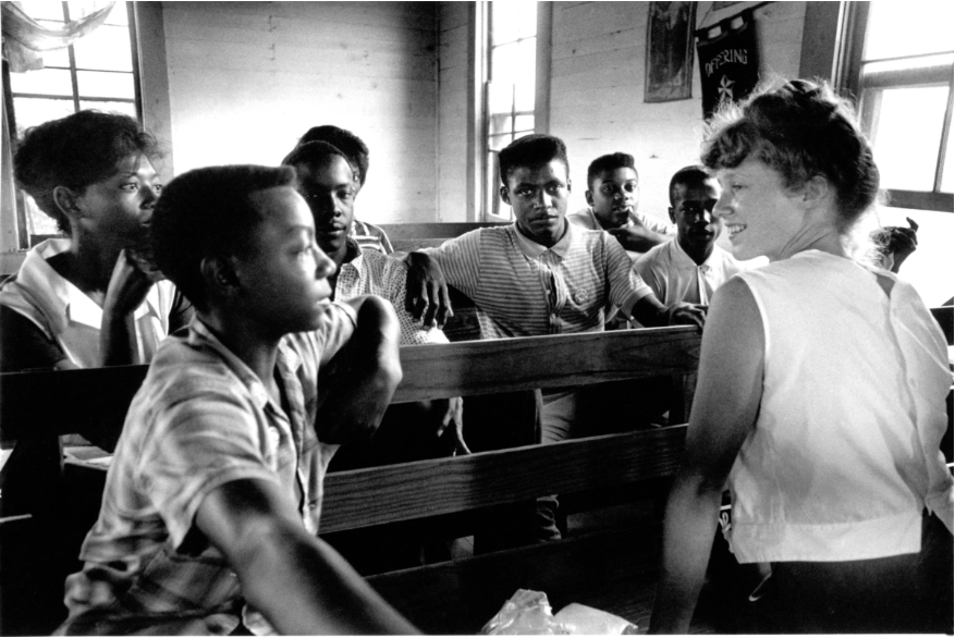 A film still from the documentary Freedom Summer of a black-and-white photograph inside MS Freedom Summer School. A group of African American boys sit in pews speaking to a white woman who sits and smiles at them.