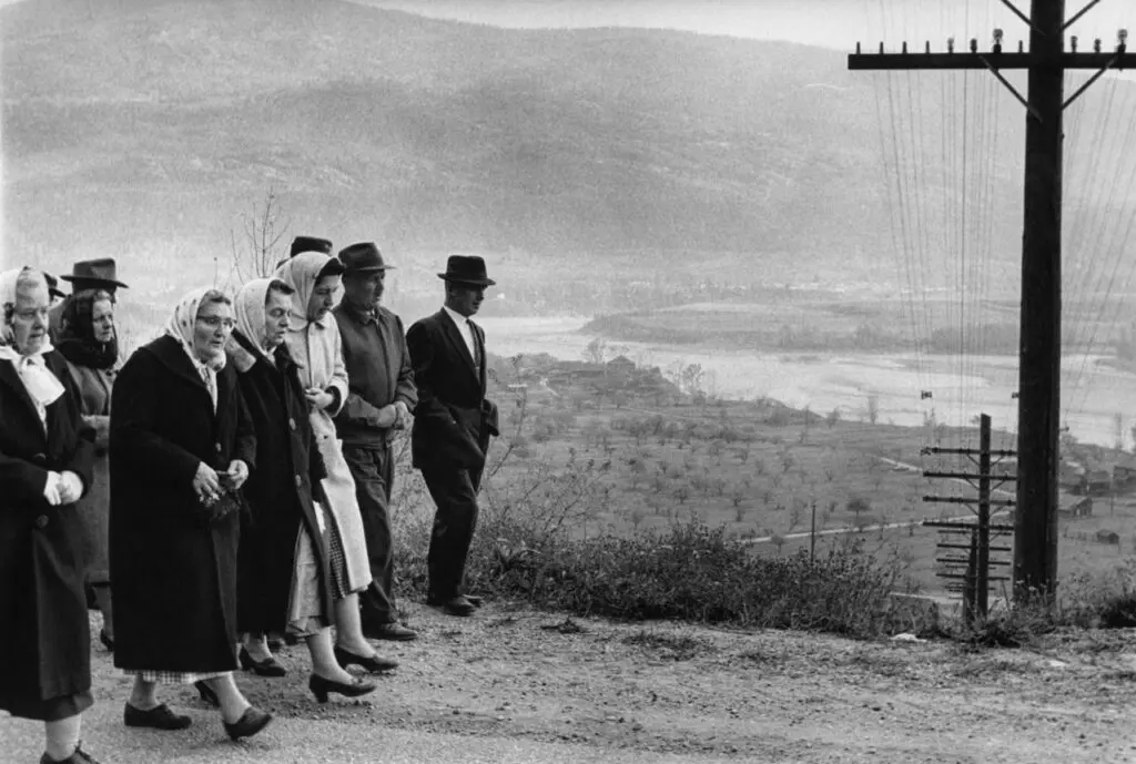 A black-and-white photograph by Tamio Wakayama of a group of elderly people walking down a dirt road, shown from the side.