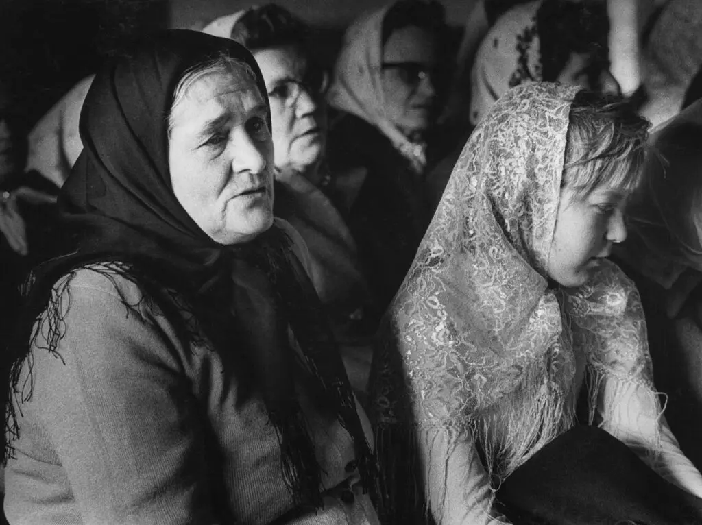 A black-and-white photograph of a middle-aged Doukhobor woman sitting in a pew next to a girl. Both women wear scarves on their heads.