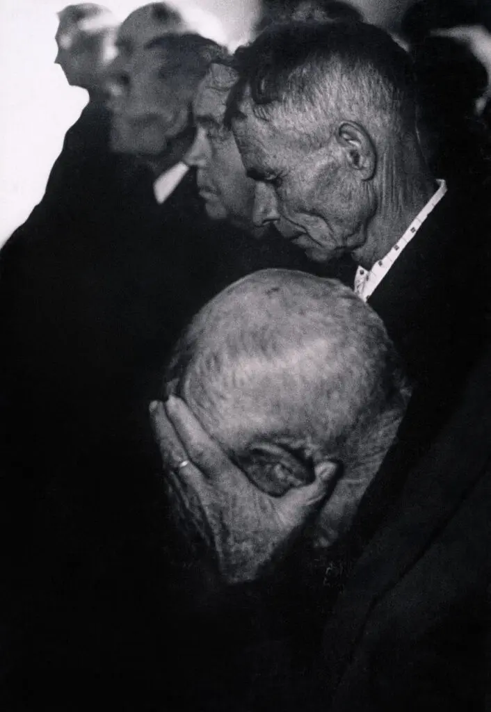 A black-and-white vertical portrait of elderly men. The man in the foreground has his head in his hands. The image has a sombre tone.
