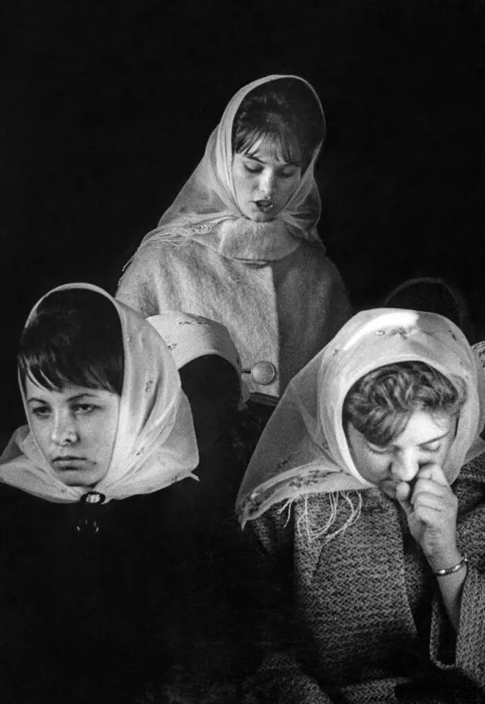 A black-and-white vertical portrait of three women, each wearing white scarves on their heads. They look sombre.