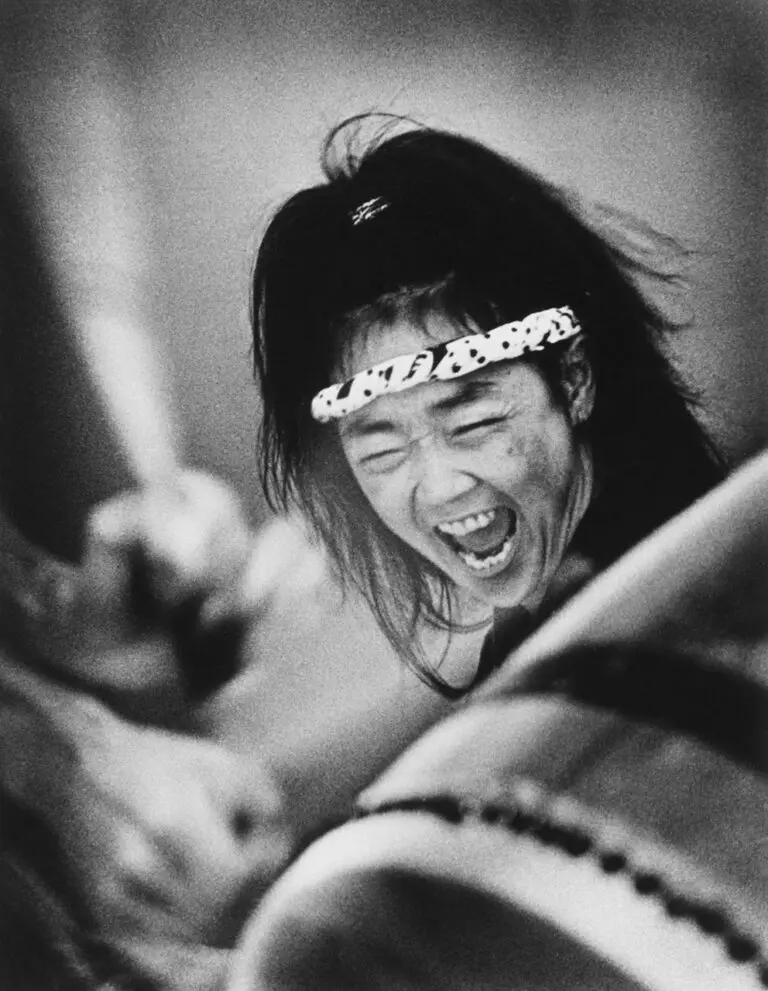 Black-and-white photograph of a close-up of woman Taiko drummer. We see her drum in the foreground, and her arm raised in the background. At the centre of the image is the woman's face, with her mouth open screaming.