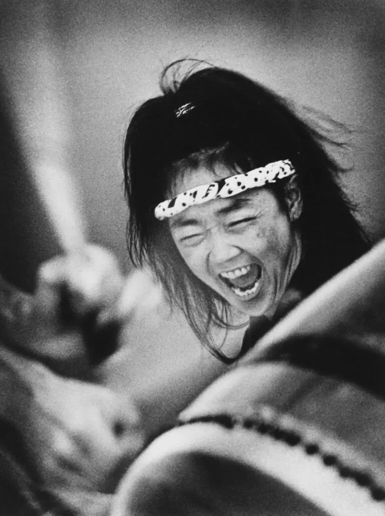 Black-and-white photograph of a close-up of woman Taiko drummer. We see her drum in the foreground, and her arm raised in the background. At the centre of the image is the woman's face, with her mouth open screaming.