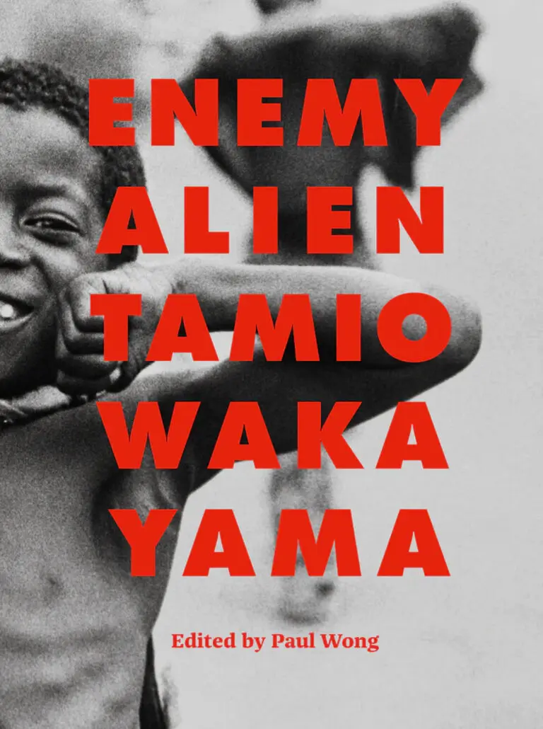 Cover of an exhibition catalogue with red, capital letters that read "Enemy Alien Tamio Wakayama." Below the title, in a smaller font, it reads: "Edited by Paul Wong." The red letters appear over a black-and-white photograph of a young, black boy with no shirt on, who smiles and beams out at the camera, while seemingly flexing his muscles.