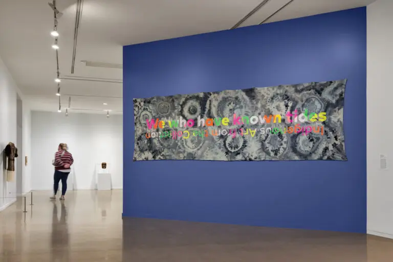 Installation photograph of the exhibition "We who have known tides: Indigenous Art from the Collection." We see a blue wall to the right of the image with an ice-dyed fabric work on it that has neon multicoloured text on it with the exhibition title. The subtitle is upside down. To the left, we see a woven jacket from the side on the wall, and a woman is slightly blurred as she enters the exhibition space.