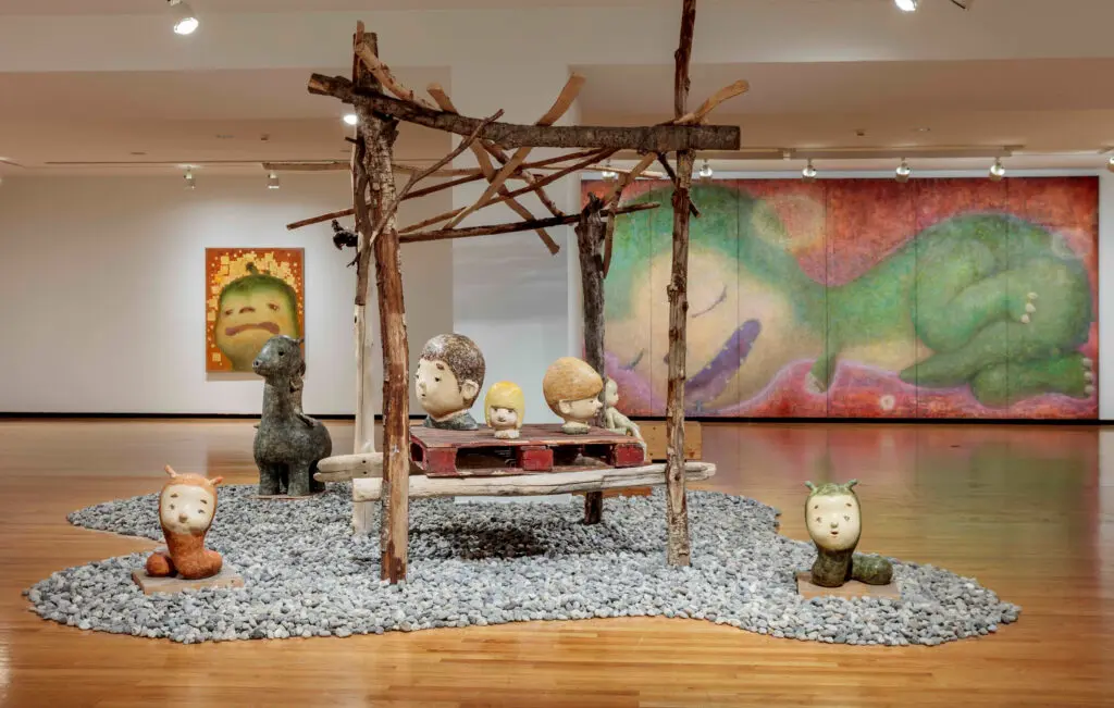 Installation photograph of Otani Workshop's exhibition "Otani Workshop: Monsters in My Head." We see what looks like an island made up of small stones in the middle of a room. There is a pagoda-like structure in the middle of it made of wooden sticks. On either side of the structure, we see two small worm- or caterpillar-looking ceramic sculptures. On the structure, there are three ceramic sculptures of heads. In the background, we see two paintings on the wall of Otani's dinosaur-like figure Tanilla.