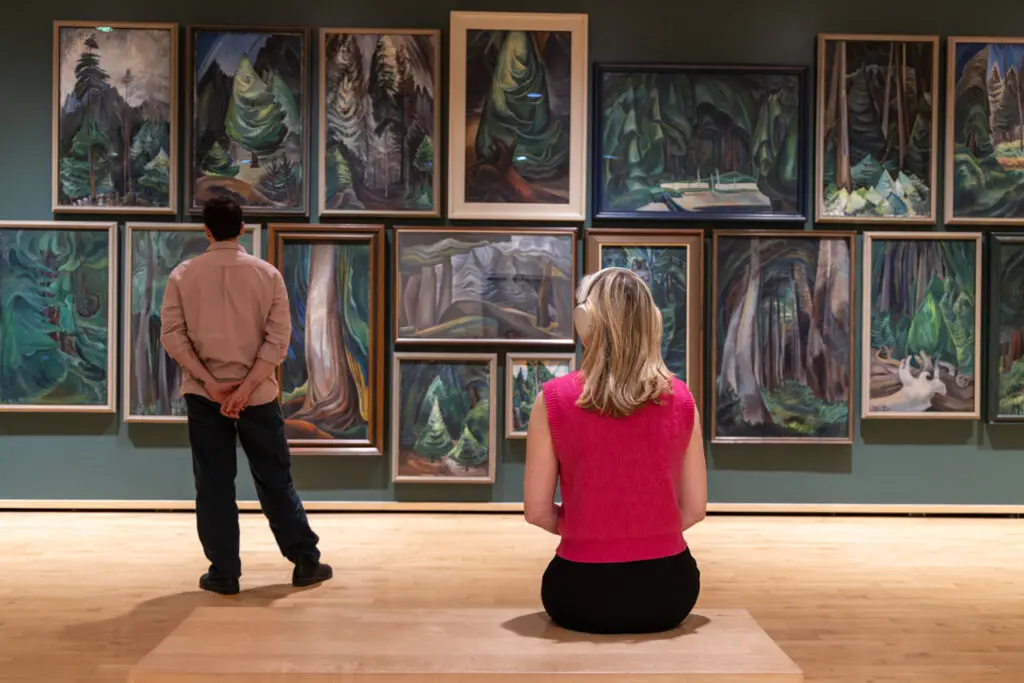 A photograph of two people looking at the forest paintings of Emily Carr. One woman sits on a bench, and a man stands closer to the wall.