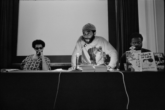 A film still from the documentary True North of a black-and-white photograph of poet and activist Ted Joans half standing and speaking into a microphone at a table with a black table cloth. He has two men sitting on either side of him. They are at the Congress of Black Writers in Montreal, Canada, 1968.