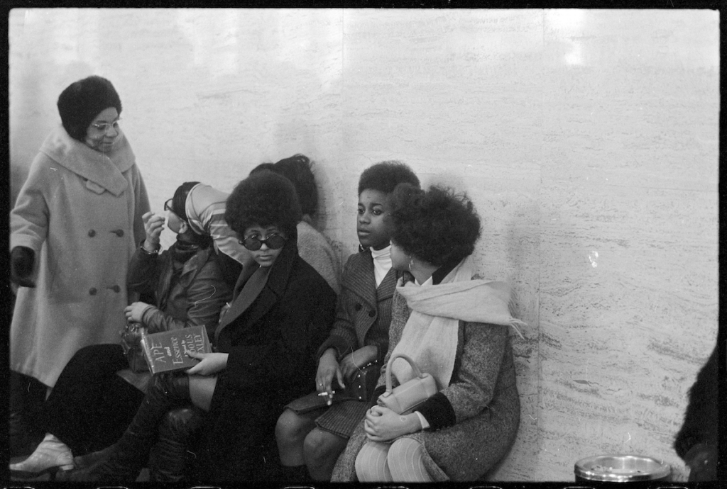 A film still from the documentary True North of a black-and-white photograph of Black women gathered outside, sitting on a bench and leaning against a blank wall.