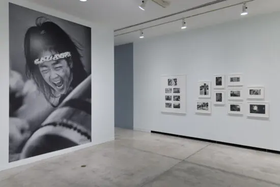 Installation view of the exhibition Enemy Alien: Tamio Wakayama. In this photograph, there are black-and-white photographs in white frames on the right wall, and on the left there is a large black-and-white mural of a close-up of woman taiko drummer. We see her drum in the foreground, and her arm raised in the background. At the centre of the image is the woman's face, with her mouth open screaming.