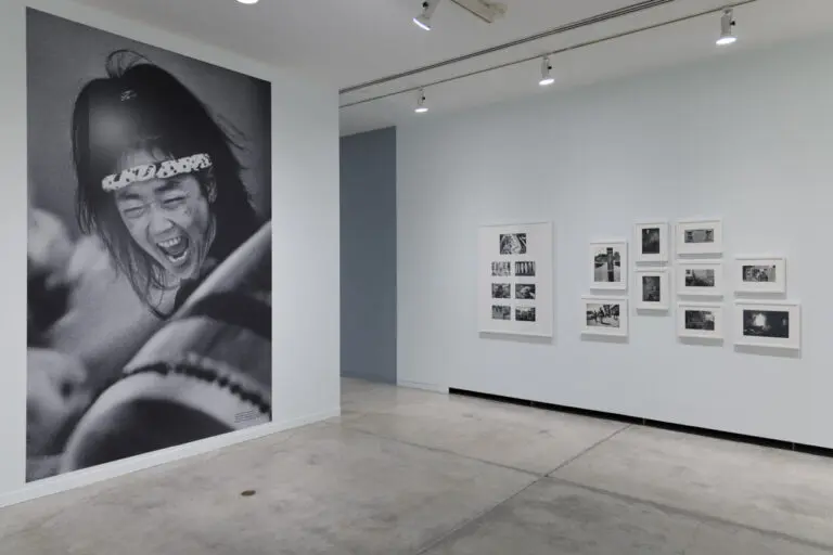 Installation view of the exhibition Enemy Alien: Tamio Wakayama. In this photograph, there are black-and-white photographs in white frames on the right wall, and on the left there is a large black-and-white mural of a close-up of woman taiko drummer. We see her drum in the foreground, and her arm raised in the background. At the centre of the image is the woman's face, with her mouth open screaming.