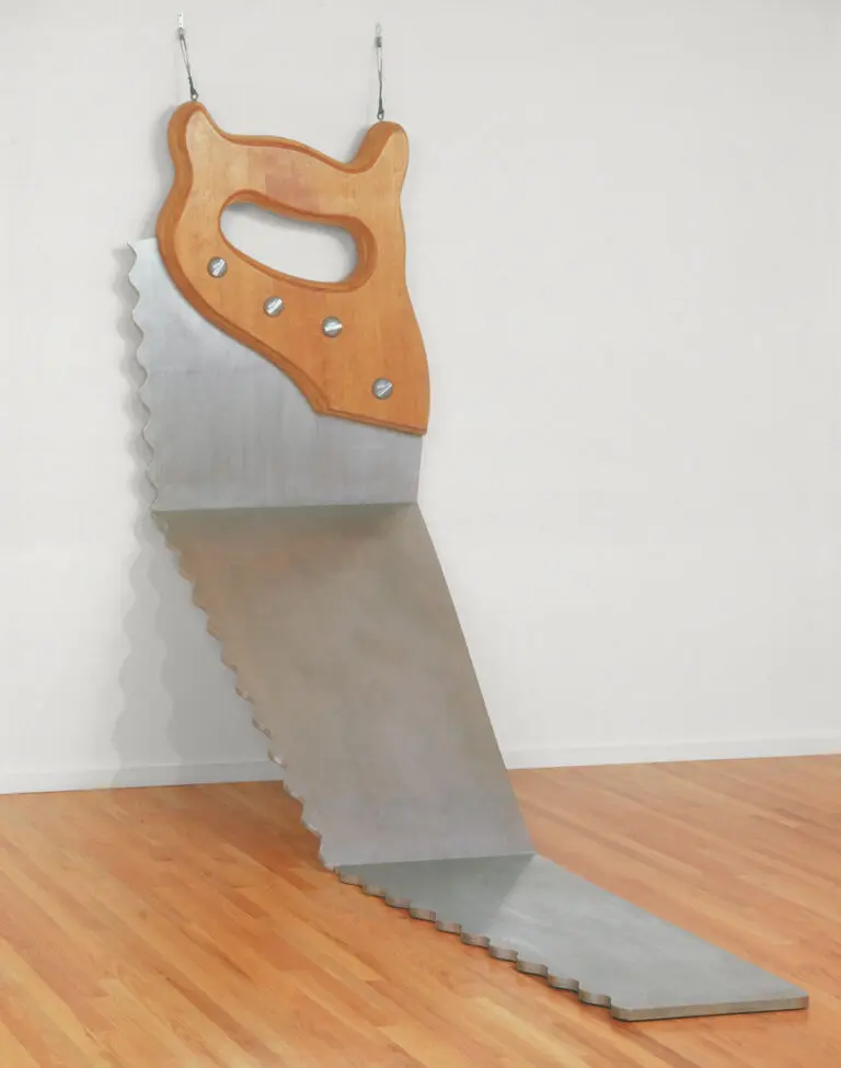 A photograph of a large, oversize saw. It leans agains a white wall. The handle leans agains the wall, and then the blade bends in two places down to the floor.