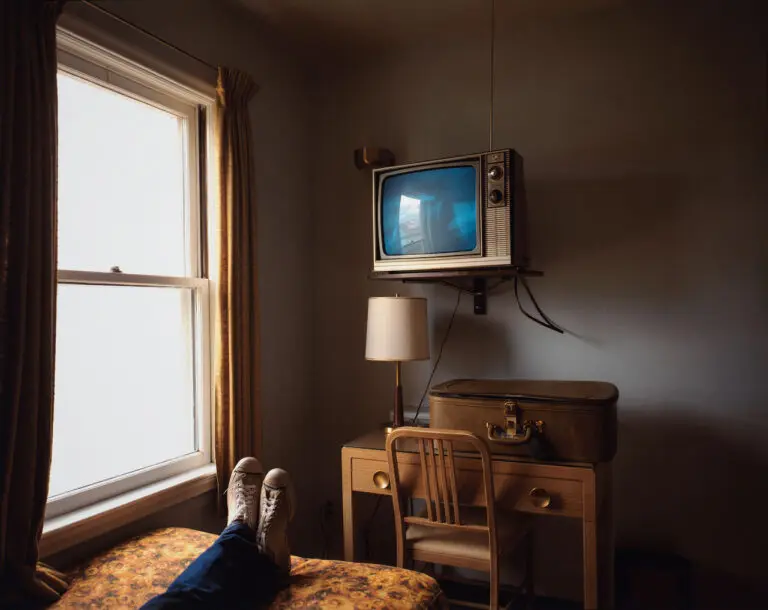 A colour photograph by Stephen Shore of a hotel room. We see a person's feet on a bed and a television set over a desk.