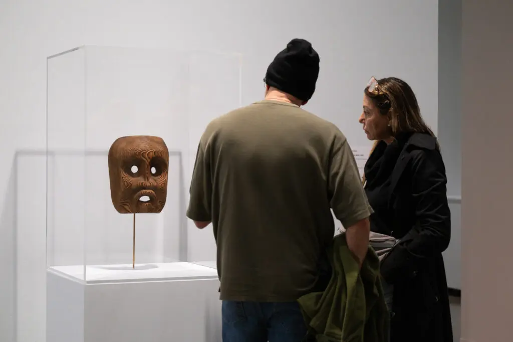 A photograph of a man and a woman from behind looking at a plinth with a carved wooden Indigenous mask in it.