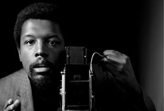 A black-and-white photograph of a Black man from the torso up standing behind a camera and looking out at the viewer. His hand is raise in the left side of the image, as if directing the scene.
