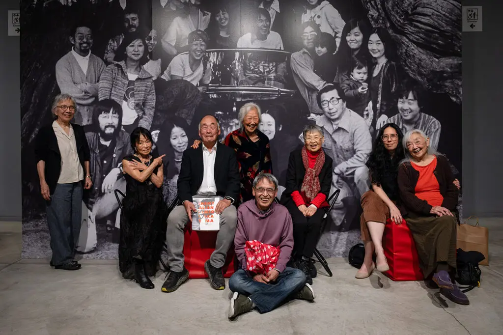 A photograph of a group of people standing and sittting in front of a black-and-white photo mural of members of the Japanese Canadian Centennial Project (JCCP) in front of a hollow tree in Stanley Park, Vancouver, British Columbia, June 1976. The people in the foreground are those in the photograph behind them in present day.
