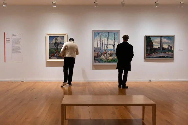 A photograph of two people looking at three paintings by Emily Carr on a white gallery wall
