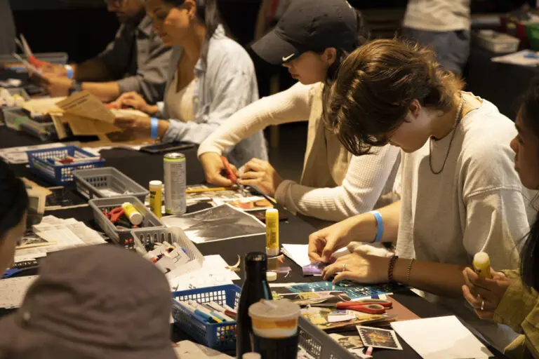 A photograph of people sitting at a table collaging–holding scissors, cutting, looking down at magazines.