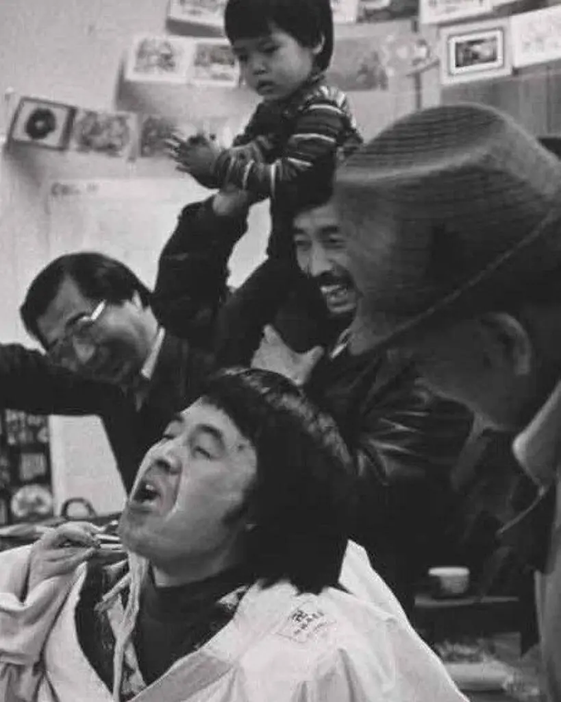Detail from a black-and-white photograph by artist Tamio Wakayama taken at a Christmas party at Tonari Gumi in Vancouver in 1978. We see people being silly, with a central figure making a face, and everyone around him laughing. A young boy sits on a man's shoulders in the background.