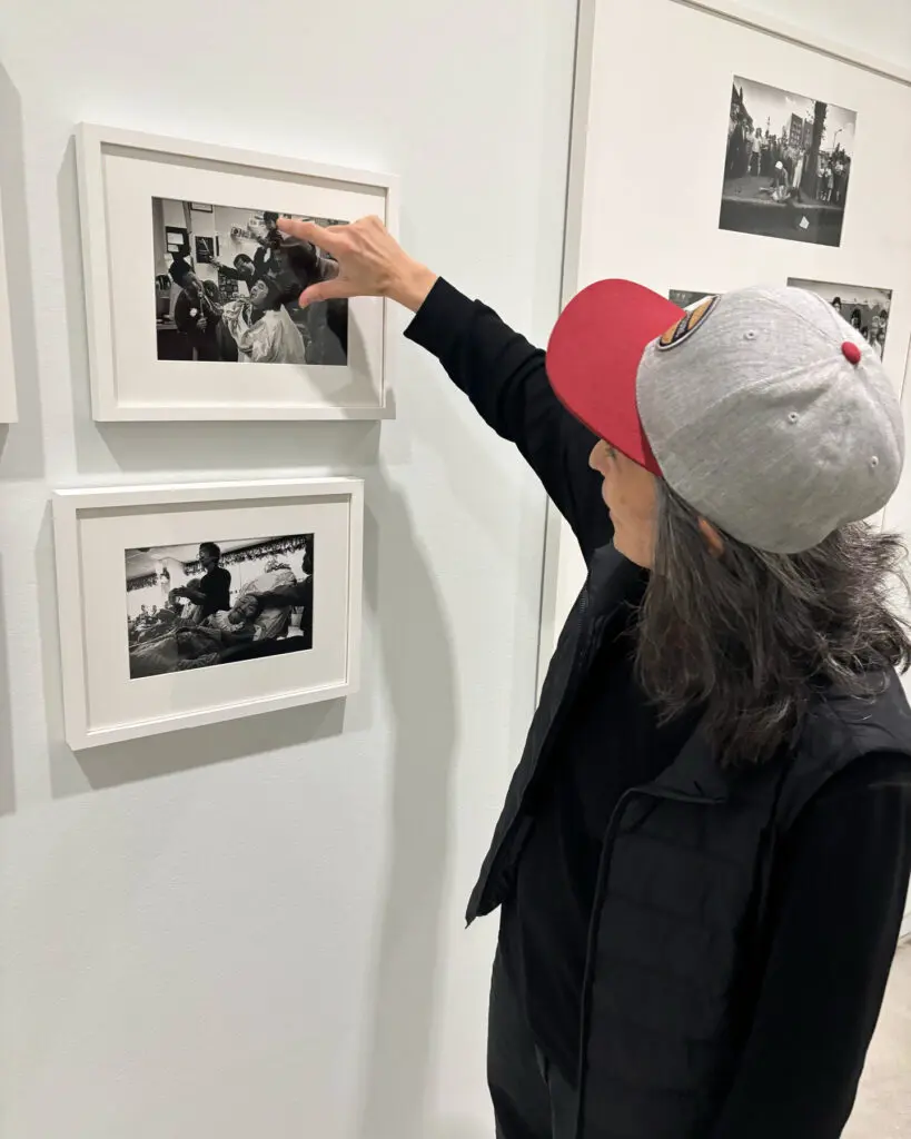 A photography of E.Kage standing in the exhibition "Enemy Alien: Tamio Wakayama" looking closely at white framed black-and-white photographs. Kage stands close to the image and points out their father in one image.