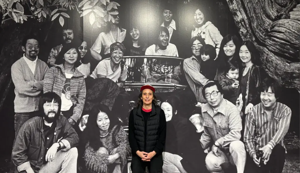 A photograph of a person standing in front of a black-and-white photo mural of members of the Japanese Canadian Centennial Project (JCCP) in front of a hollow tree in Stanley Park, Vancouver, British Columbia, June 1976