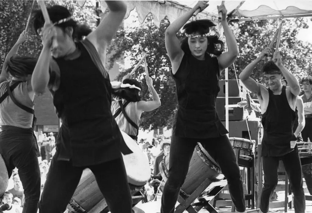 A black-and-white photograph by artist Tamio Wakayama of taiko drumming performers, including E.Kage