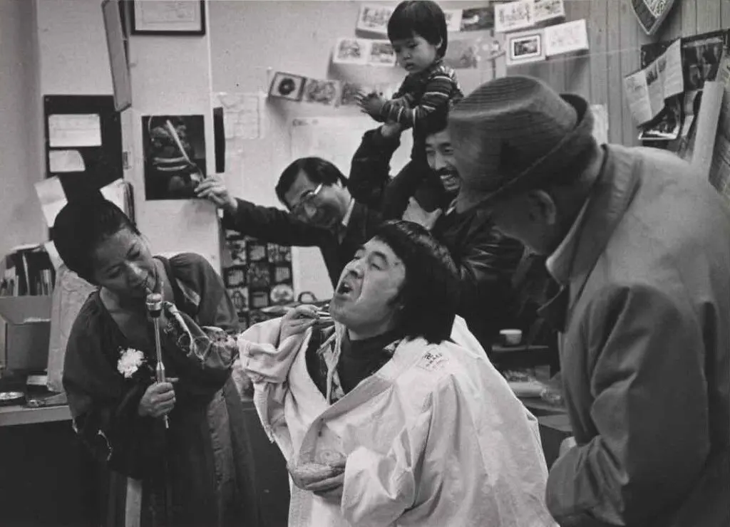 A black-and-white photograph by artist Tamio Wakayama taken at a Christmas party at Tonari Gumi in Vancouver in 1978. We see people being silly, with a central figure making a face, and everyone around him laughing. A young boy sits on a man's shoulders in the background.