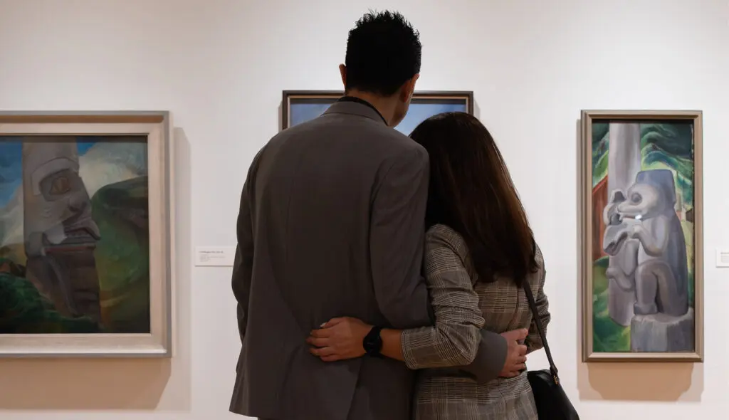 A photograph of a couple, a man and a woman, fro.m behind, embracing each other as they look at paintings by Emily Carr on gallery walls.
