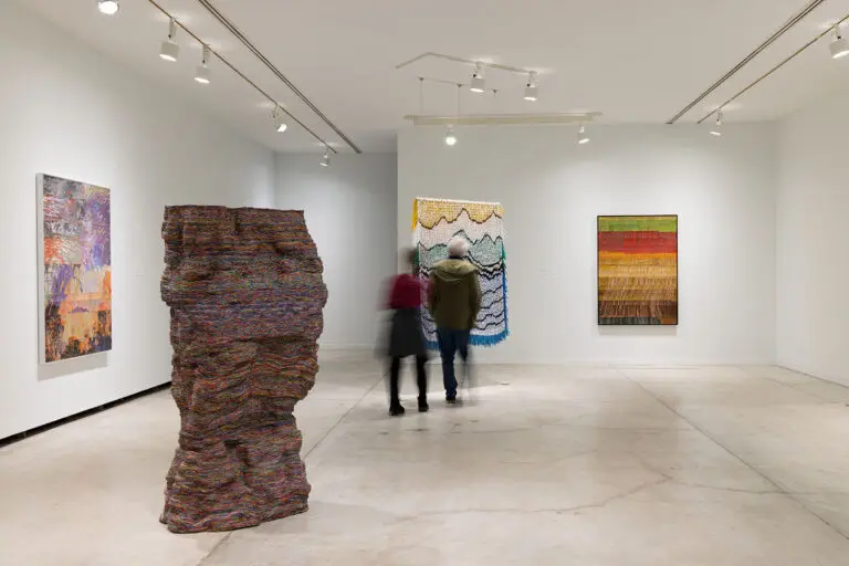 Installation view of 4 artworks by Emily Hermant in the exhibition Vancouver Now!. At the centre of the image, a couple, a man and a woman, look at the back of an hanging, tapestry-like work.