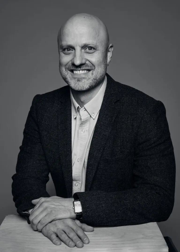 A black-and-white headshot of Grant Mosby, a white man in a black suit jacket, seen from the waist up. His hands are out in front of him.