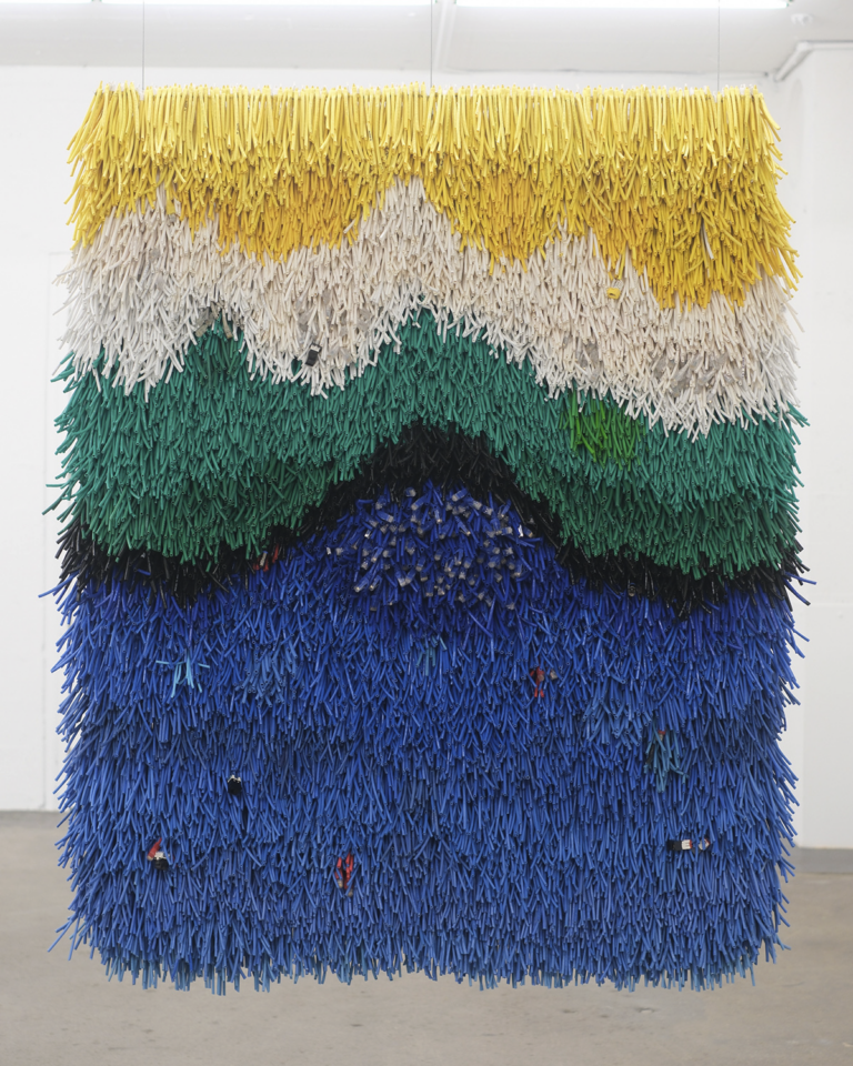 A photography of a weaving work by Emily Hermant seen from the front. The weaving, which has a fringe on the front, evokes a landscape made up of rows of colour. From top to bottom, we see yellow, white, greens, dark blue, and then blues. At the centre, we see some additional objects incorporated.