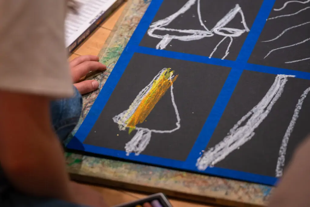 A photograph of a child's pastel drawing of trees in white on black paper. We see a corner of the paper and a child's arm in the left of the image.