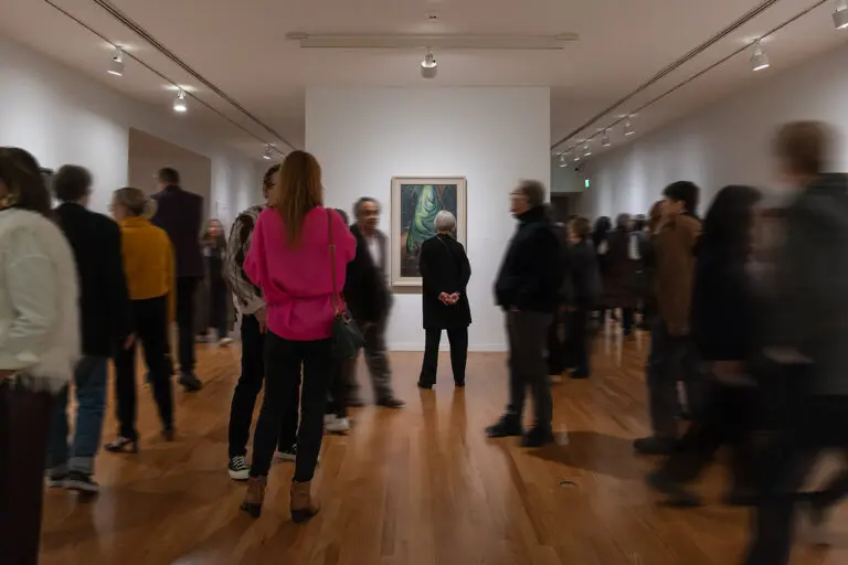 A photograph of a room full of people enjoying art together. At the centre, a woman stands and looks at a painting of a tree by Emily Carr.