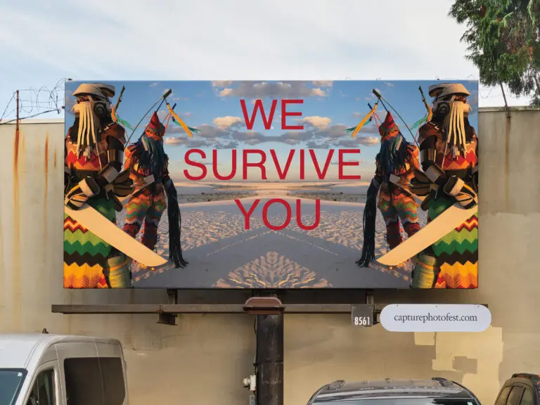 A mock-up image of a billboard outside in a parking lot. The billboard features an artwork by Cannupa Hanska Luger with 4 people, 2 on either side of the billboard, dressed in futuristic looking traditional Indigenous clothing, and in the middle the text "WE SURVIVE YOU" In red all caps.