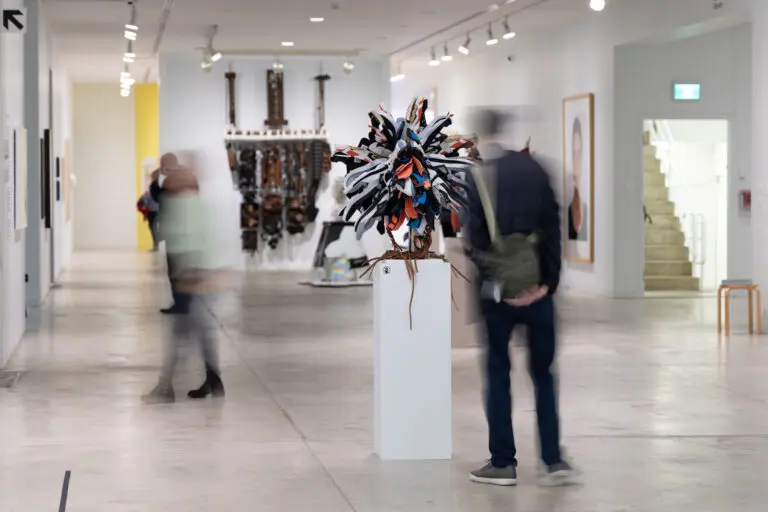A photograph of two blurred people looking at the exhibition Highlights from the Collection. One man in the foreground looks at a sculptural work by Brian Jungen made up of sneakers, and in the back we see a large, brown structure that looks like a de-constructed piano hanging on the wall.