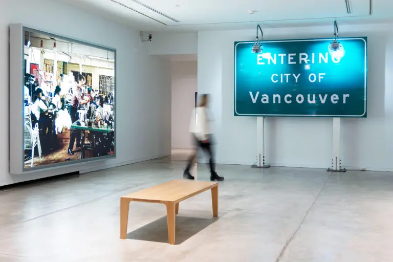 A photograph of the exhibition Highlights from the Collection, featuring a light box work by Jeff Wall and a green highway sign with lights on top that reads: Entering the City of Vancouver, but Ron Terada. A woman is blurred at the centre of the image as she walks through the room.